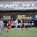 Players engage in a competitive soccer game on a field while others are seated on the bench watching attentively. The action is taking place in front of a wooden structure displaying multiple advertisements, including orthodontics, lacrosse, and field hockey.