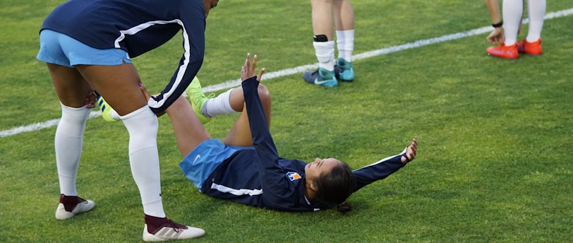A group of athletes in sportswear are on a green soccer field. Two individuals are interacting closely; one helps the other who is lying on the grass. Nearby, other athletes are stretching.