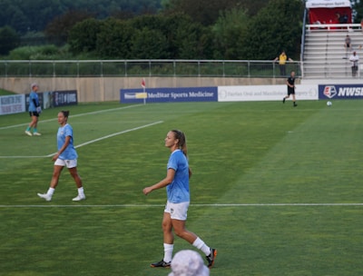 A soccer game scene on a green grass field with players wearing light blue jerseys and white shorts. A goalpost is visible in the background, and there are people sitting or standing on the bleachers. Advertising banners are displayed along the sidelines.