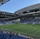 A modern sports stadium with a large, green soccer field and surrounding seating areas. Blue and yellow seats are visible, with some spectators scattered across the stands. The stadium is partially covered with a steel lattice roof, showing a clear blue sky. Several security personnel in high-visibility vests are present.