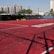 A sports court with a red playing surface and white lines, surrounded by a black mesh fence. In the background, tall buildings are visible, along with some trees and a few yellow chairs positioned near the fence.